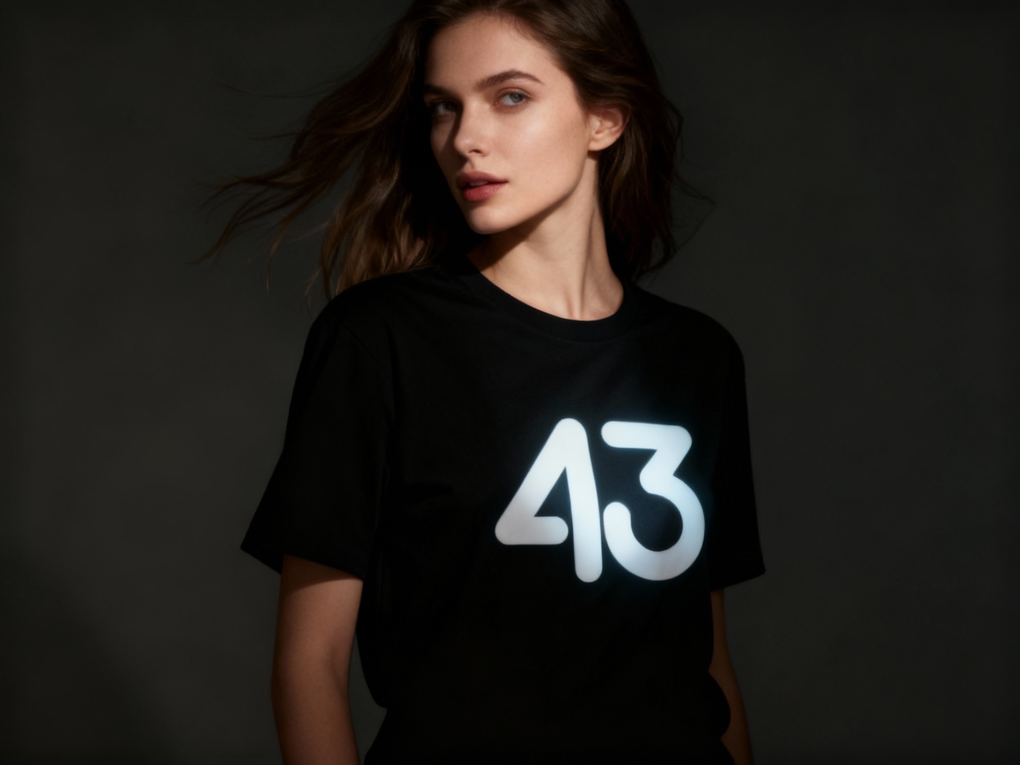 提取参考图的LOGO单独呈现，改为白色，25岁欧洲女性模特，身着黑色丝光棉T恤，白色LOGO以UV凸印工艺呈现，触感细腻有轻微立体感，侧光拍摄，光线照亮LOGO部分，与T恤暗部形成对比，模特转身回眸，发丝飘动，背景为低饱和暗色调，突出动态与LOGO焦点