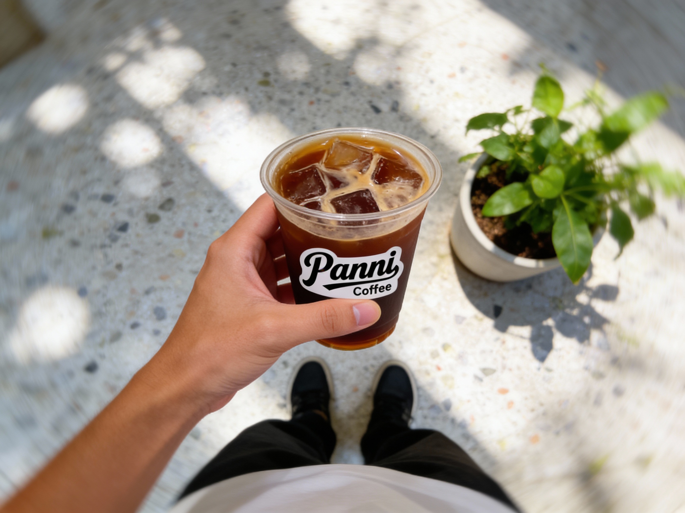 设计社交分享场景图（POV视角）。第一人称视角低头看，一只手拿着带有（智能提取参考图中的LOGO主体并去除原背景）的冰美式，LOGO清晰且无背景色残留。脚下是光影斑驳的水磨石地面或木地板，旁边是好看的绿植盆栽。画面动态模糊，色彩清新，高光柔和，极具生活气息和打卡欲望。