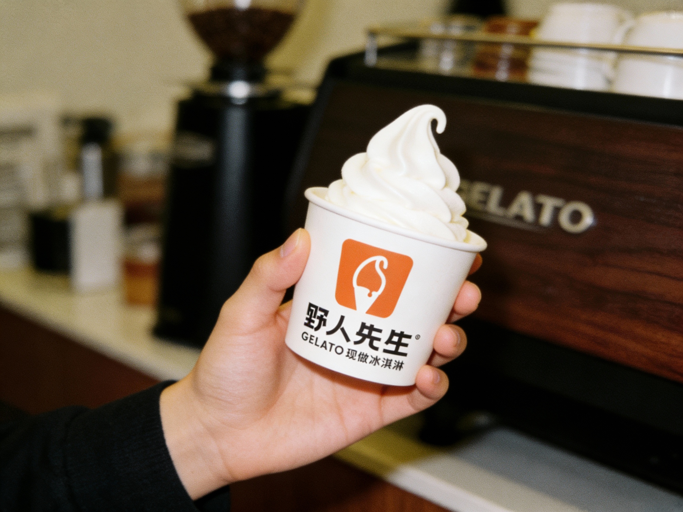 一张极具'网感'的顾客社交分享抓拍图（UGC风格）。画面是一名顾客的手部特写，手持一杯印有（将参考图中的LOGO）的冰淇淋，背景是虚化处理的咖啡厅一角，隐约可见装修中的黑胡桃木质感或品牌物件。构图采用非对称、略微倾斜的角度，带有轻微的动态模糊，模拟胶片相机的颗粒感和低光环境下的氛围感。光影斑驳陆离，色彩自然复古，传达出'我在喝这家很酷的冰淇淋'的生活方式感。