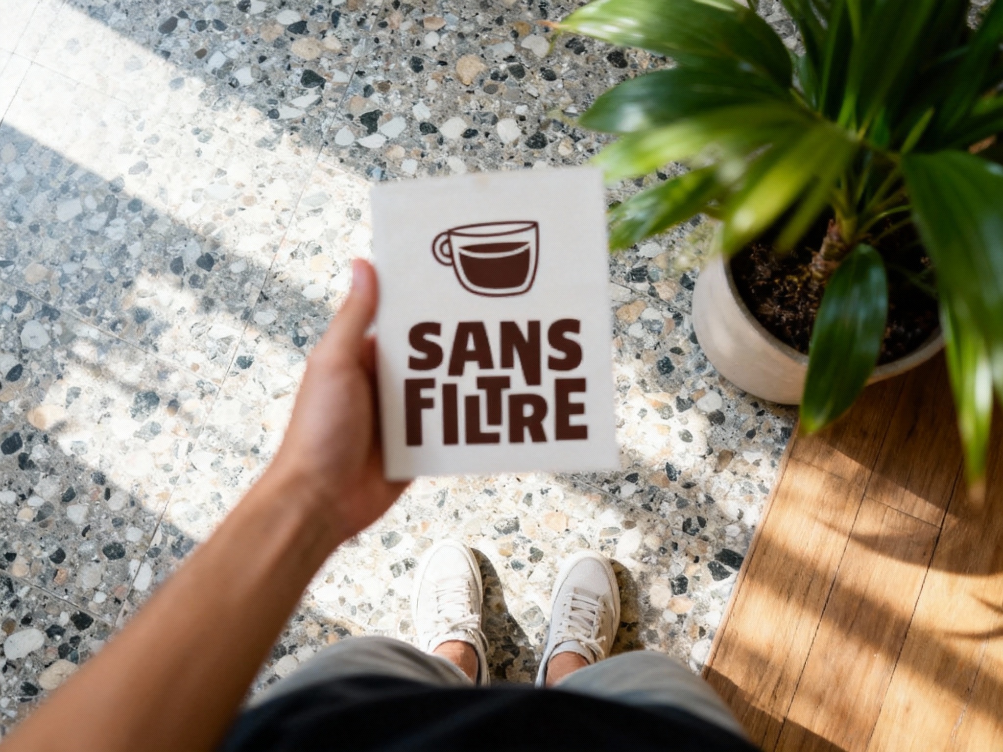 设计社交分享场景图（POV视角）。第一人称视角低头看，一只手拿着带有（智能提取参考图中的LOGO主体并去除原背景）的冰美式，LOGO清晰且无背景色残留。脚下是光影斑驳的水磨石地面或木地板，旁边是好看的绿植盆栽。画面动态模糊，色彩清新，高光柔和，极具生活气息和打卡欲望。