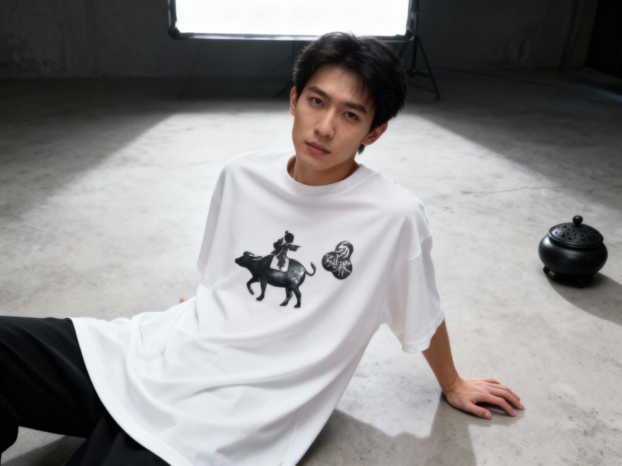 提取参考图的LOGO单独呈现，改为黑色，25岁亚洲男性模特，穿纯白oversizeT恤，黑色LOGO采用烫银工艺（呈现哑光金属质感），俯拍视角，模特单手撑地，T恤自然垂坠，LOGO位于画面中心位置，顶部柔光箱打光，消除多余阴影，背景为极简水泥灰，画面右侧放置一个黑色陶瓷香炉，与LOGO颜色一致，工业风与高级感融合