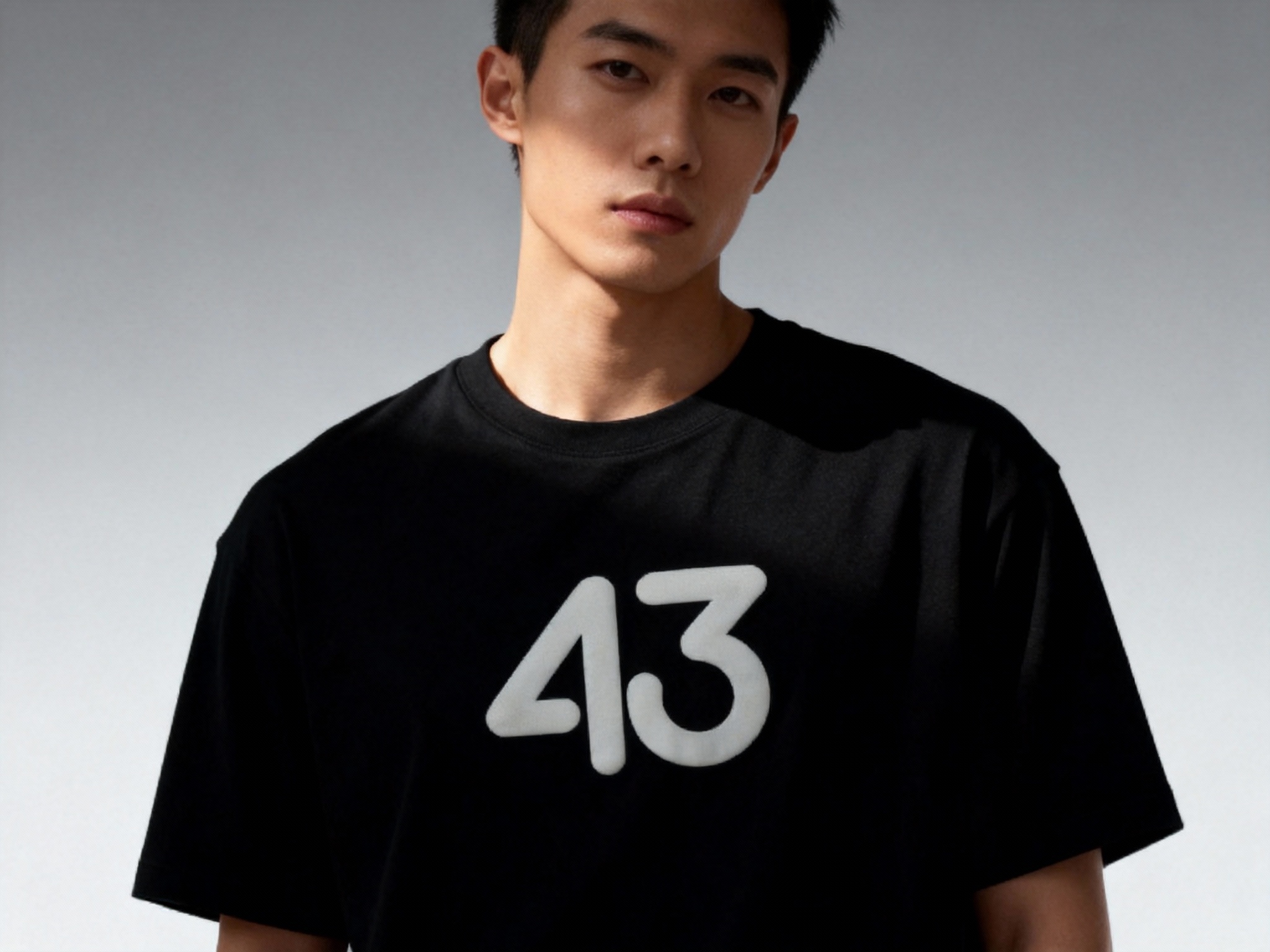 提取参考图的LOGO单独呈现，改为白色，25岁亚洲男性模特，身着纯黑重磅T恤，白色LOGO以胶浆印花工艺呈现在胸口中央，边缘平整细腻，侧逆光拍摄，光线勾勒模特肩部线条，T恤质感柔软垂顺，背景为极简灰色渐变，4A广告大片质感，视觉焦点集中于LOGO