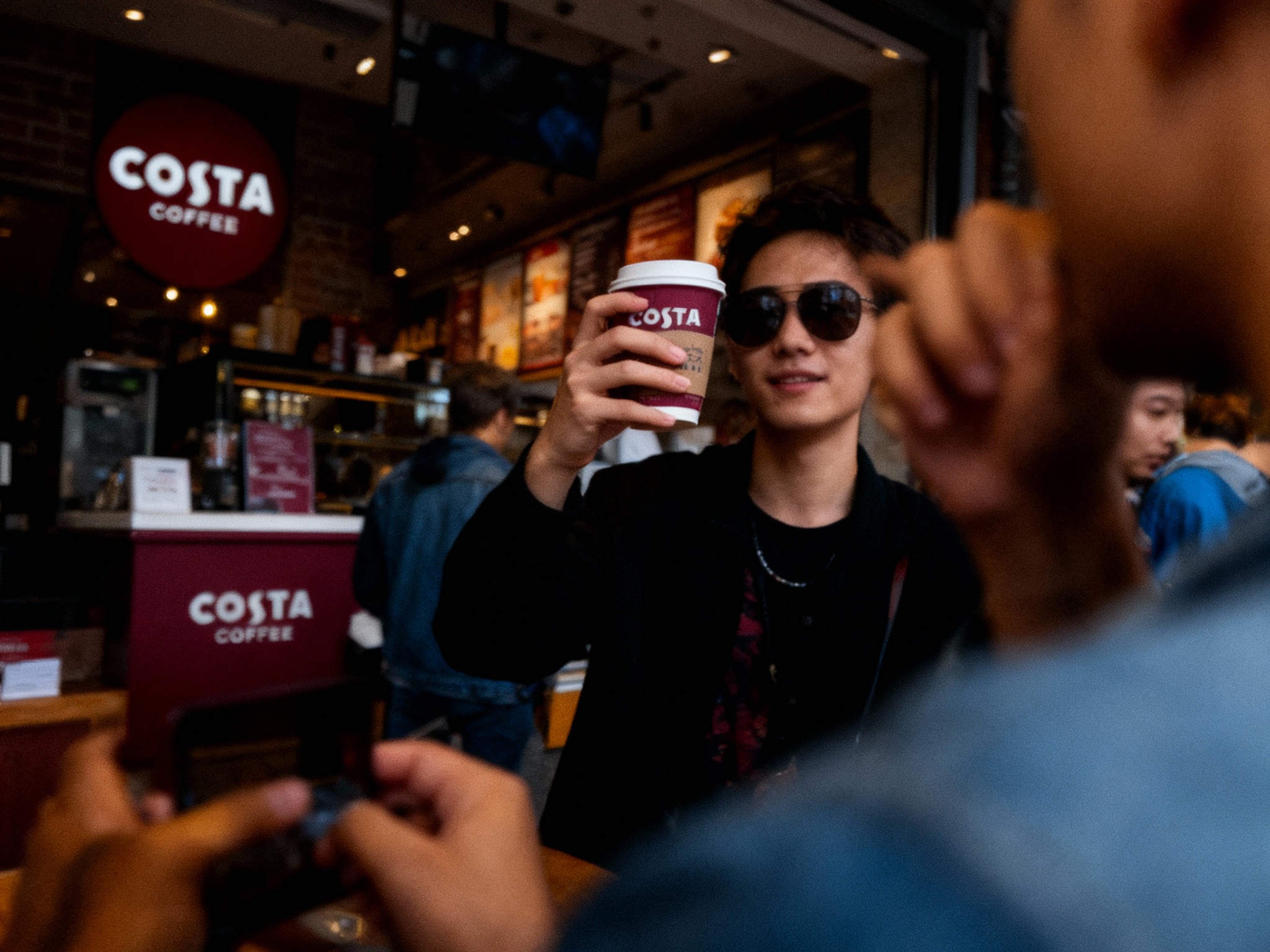 设计社交分享场景图，顾客手持带有（将参考图中的LOGO）的咖啡拍照，咖啡厅场景中（将参考图中的LOGO）融入装修或物件，场景抓拍，独特的拍摄角度，非对称，动态模糊，低光环境，意境，自然色彩，丰富层次感，细腻画质，真实，光影变幻，虚化柔和过渡，胶片质感，电影质感，大师作品，光影自然，突出品牌时尚感和年轻气质。