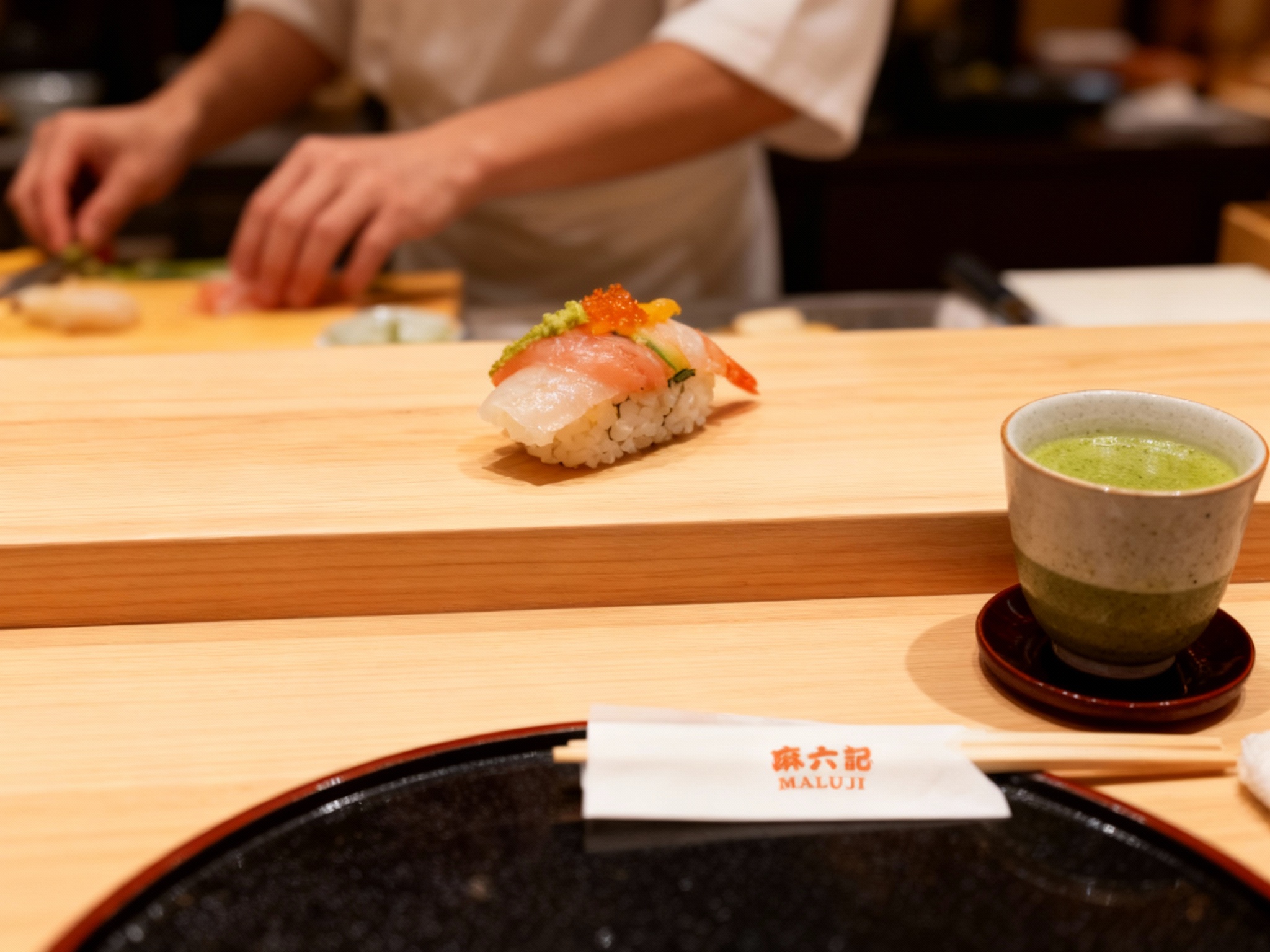 设计社交分享场景图（POV视角）。第一人称视角低头看，面前的桧木吧台上放着一贯精致的寿司或一杯抹茶，餐具上印有微小的（智能提取参考图中的LOGO主体并去除原背景）。背景虚化，可以看到厨师忙碌的手部动作。画面色彩还原度高，光泽感强，极具食欲和临场感。