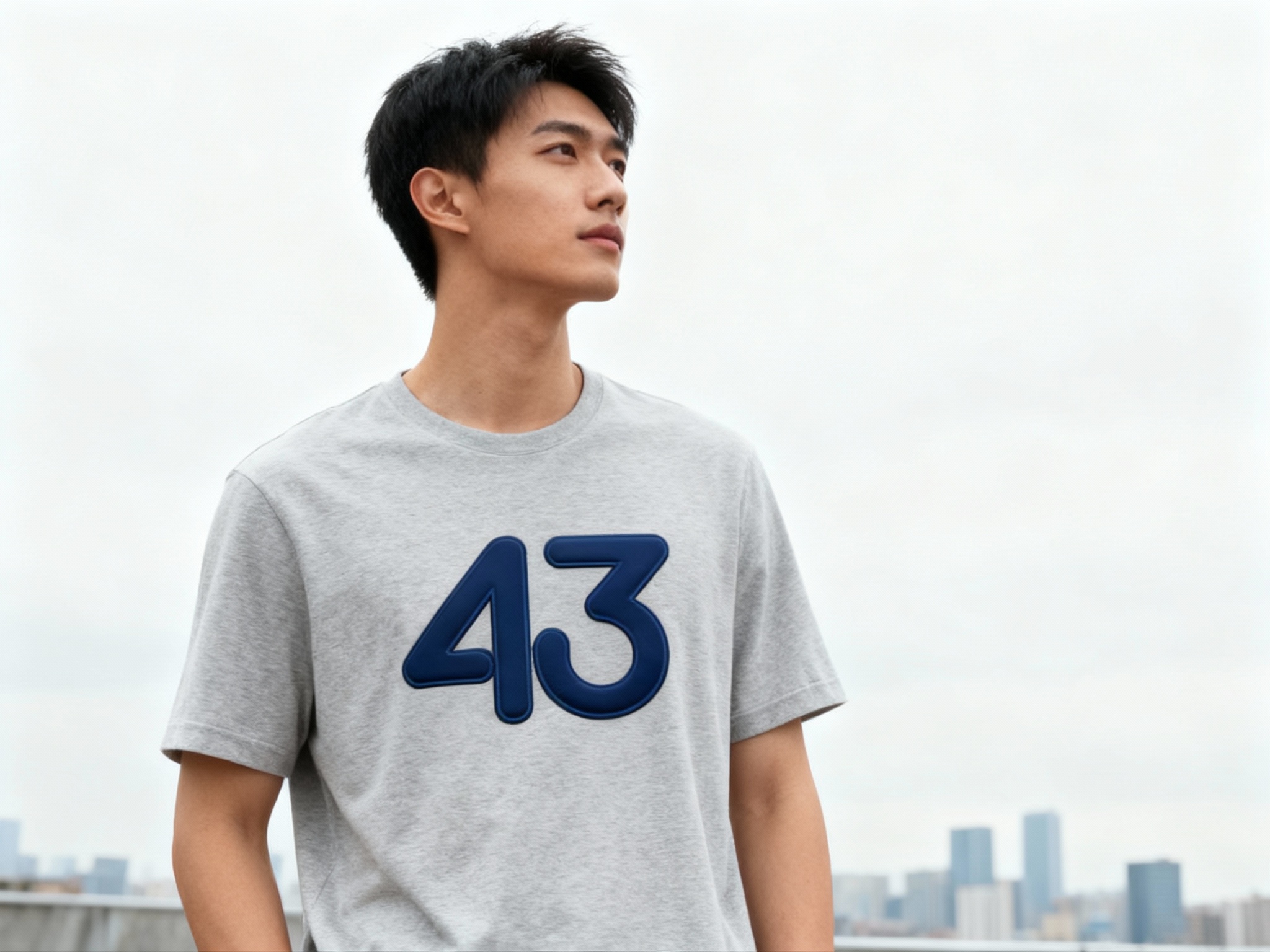 提取参考图的LOGO单独呈现，改为藏蓝色，25岁亚洲男性模特，穿浅灰纯色T恤，藏蓝色LOGO采用撞色胶浆印花，主色与T恤形成和谐对比，户外阴天拍摄，自然漫射光均匀覆盖，T恤面料纹理清晰可见，LOGO色彩饱满不褪色，模特站立远眺，背景为简洁城市天际线，大气开阔
