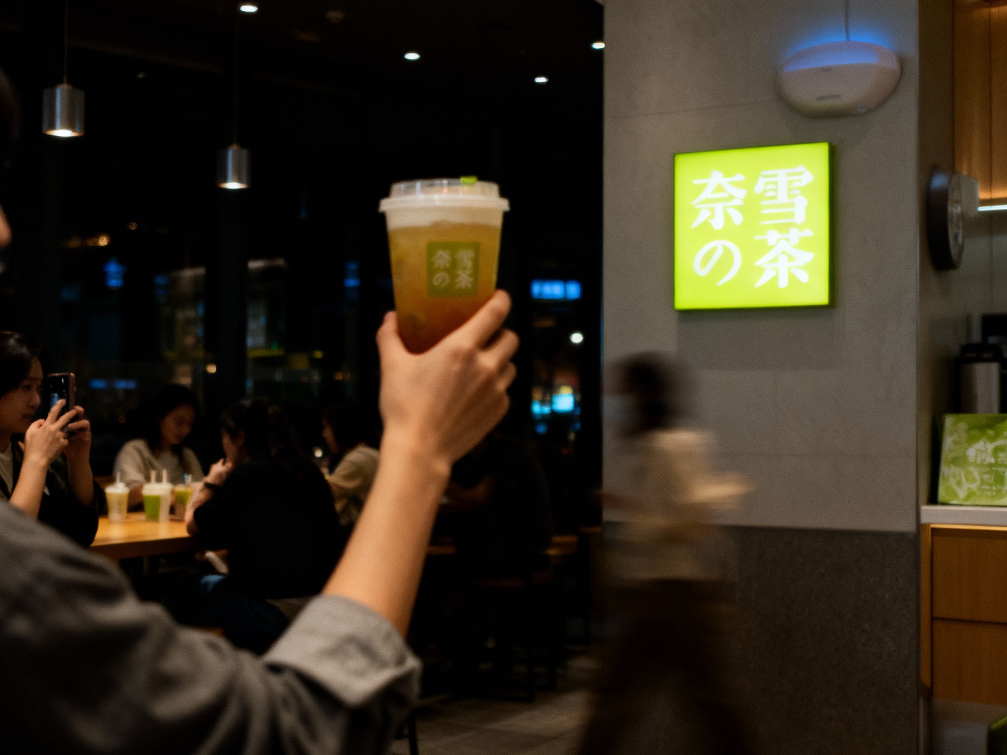 设计社交分享场景图，顾客手持带有（将参考图中的LOGO）的茶杯拍照，咖啡厅场景中（将参考图中的LOGO）融入装修或物件，场景抓拍，独特的拍摄角度，非对称，动态模糊，低光环境，意境，自然色彩，丰富层次感，细腻画质，真实，光影变幻，虚化柔和过渡，胶片质感，电影质感，大师作品，光影自然，突出品牌时尚感和年轻气质。