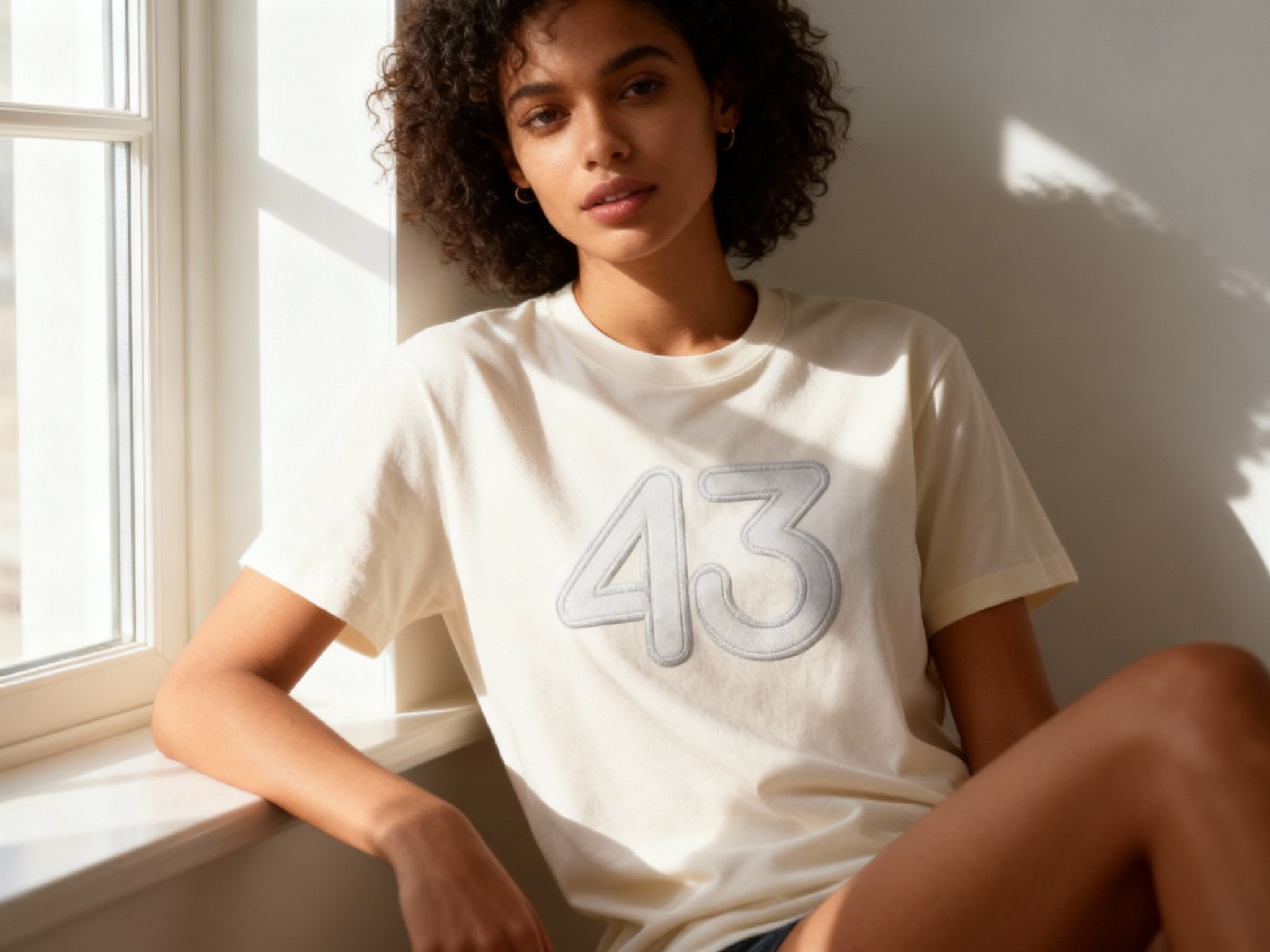 提取参考图的LOGO单独呈现，改为浅灰色，25岁混血女性模特，身着米白纯色T恤，浅灰色LOGO以丝网印刷工艺印制，色调柔和协调，窗边自然光拍摄，光线从左侧射入，在T恤表面形成明暗过渡，LOGO边缘清晰无晕染，模特靠坐窗边，姿态松弛自然，画面清新又高级