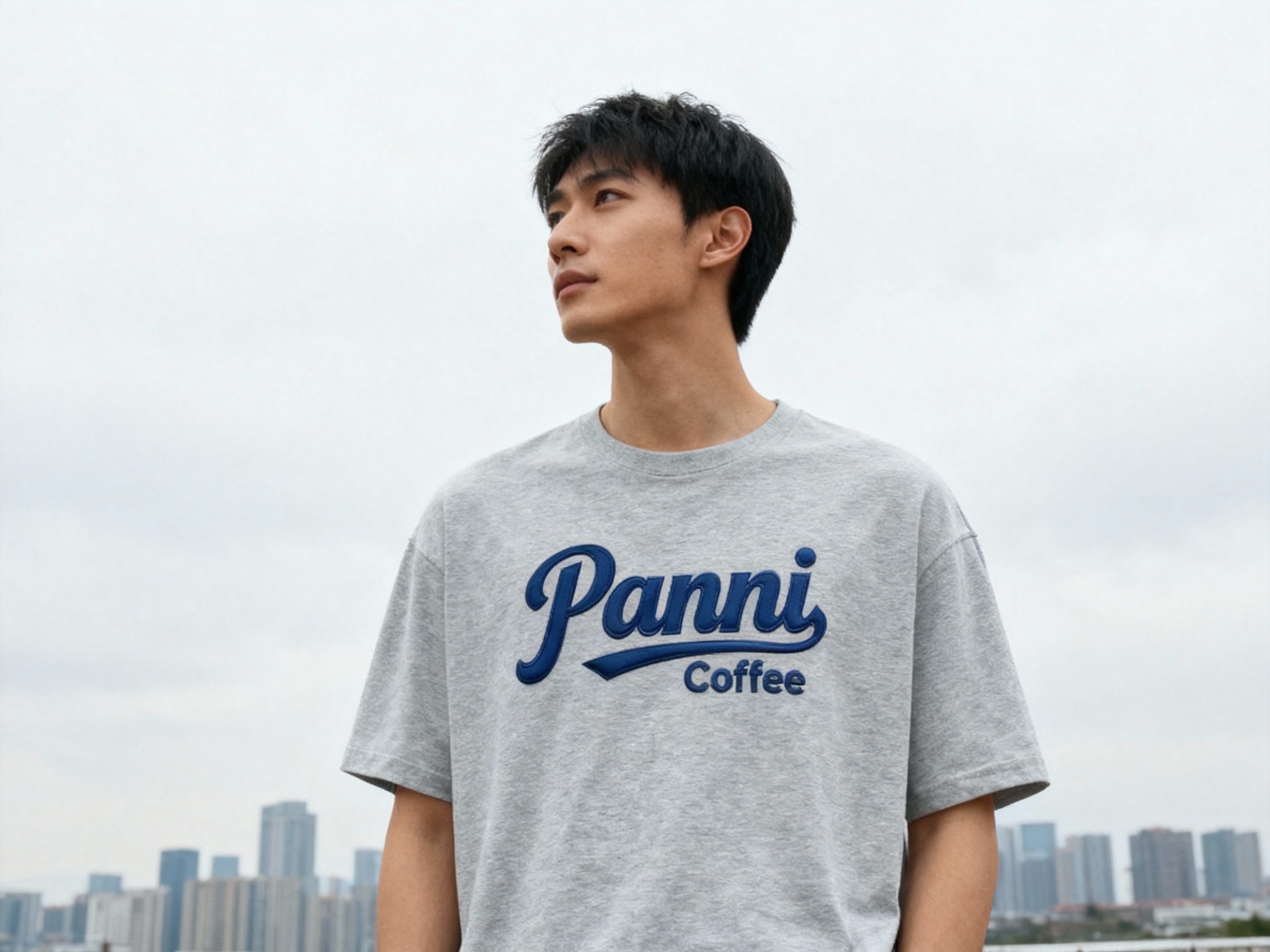 提取参考图的LOGO单独呈现，改为藏蓝色，25岁亚洲男性模特，穿浅灰纯色T恤，藏蓝色LOGO采用撞色胶浆印花，主色与T恤形成和谐对比，户外阴天拍摄，自然漫射光均匀覆盖，T恤面料纹理清晰可见，LOGO色彩饱满不褪色，模特站立远眺，背景为简洁城市天际线，大气开阔