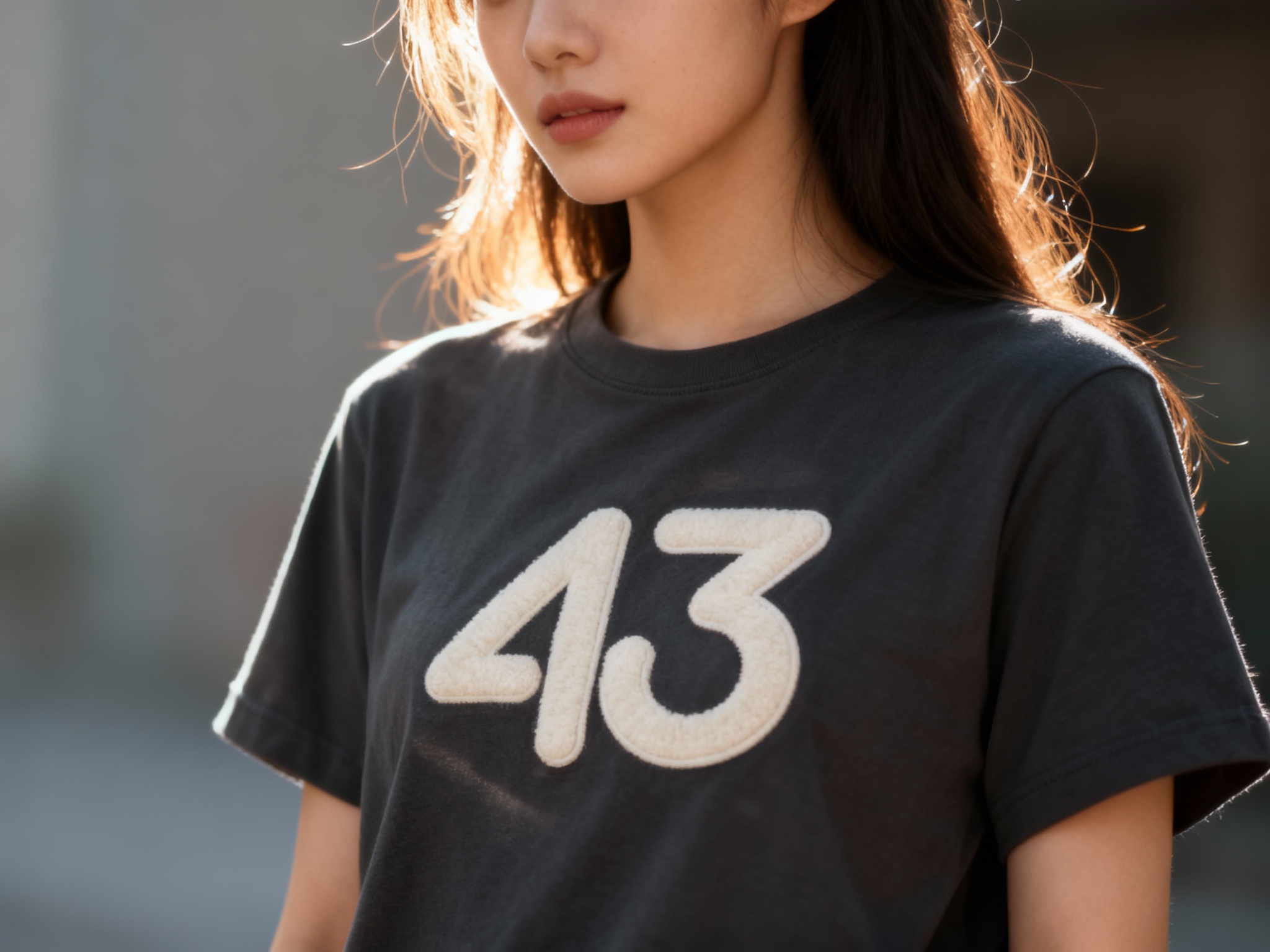 提取参考图的LOGO单独呈现，改为米白色，25岁亚洲女性模特，身着深灰纯色T恤，米白色LOGO以植绒印花工艺呈现，触感立体柔软，逆光侧拍，光线透过发丝与T恤边缘，形成柔和光晕，LOGO在侧光下呈现轻微阴影，增强层次感，背景虚化呈低饱和色调，电影感十足